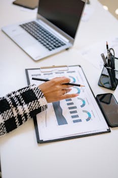 Close-up of a business setting with charts, a laptop, and a smartphone, featuring a woman's hand.